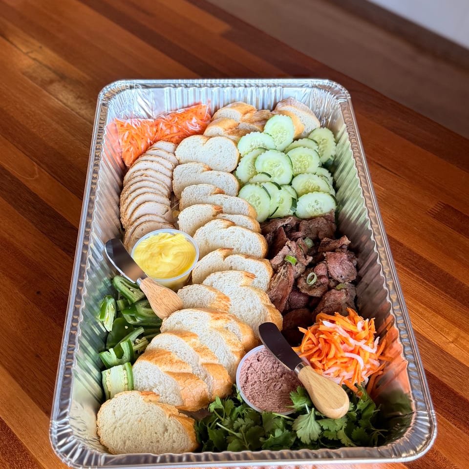 A catering tray with sliced baguettes, meats, cucumbers, pickled carrots, jalapeños, and sauces.