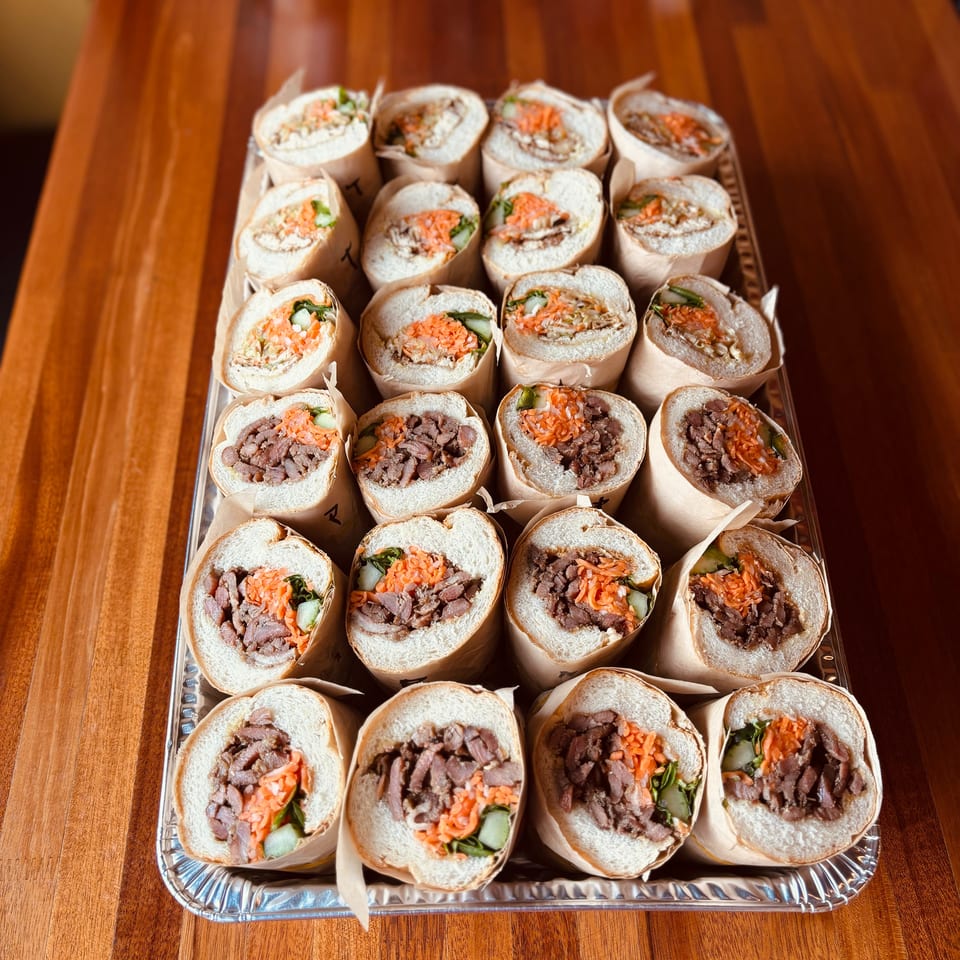 A tray of banh mi sandwiches with meat, pickled carrots, and cucumbers on a wooden table.