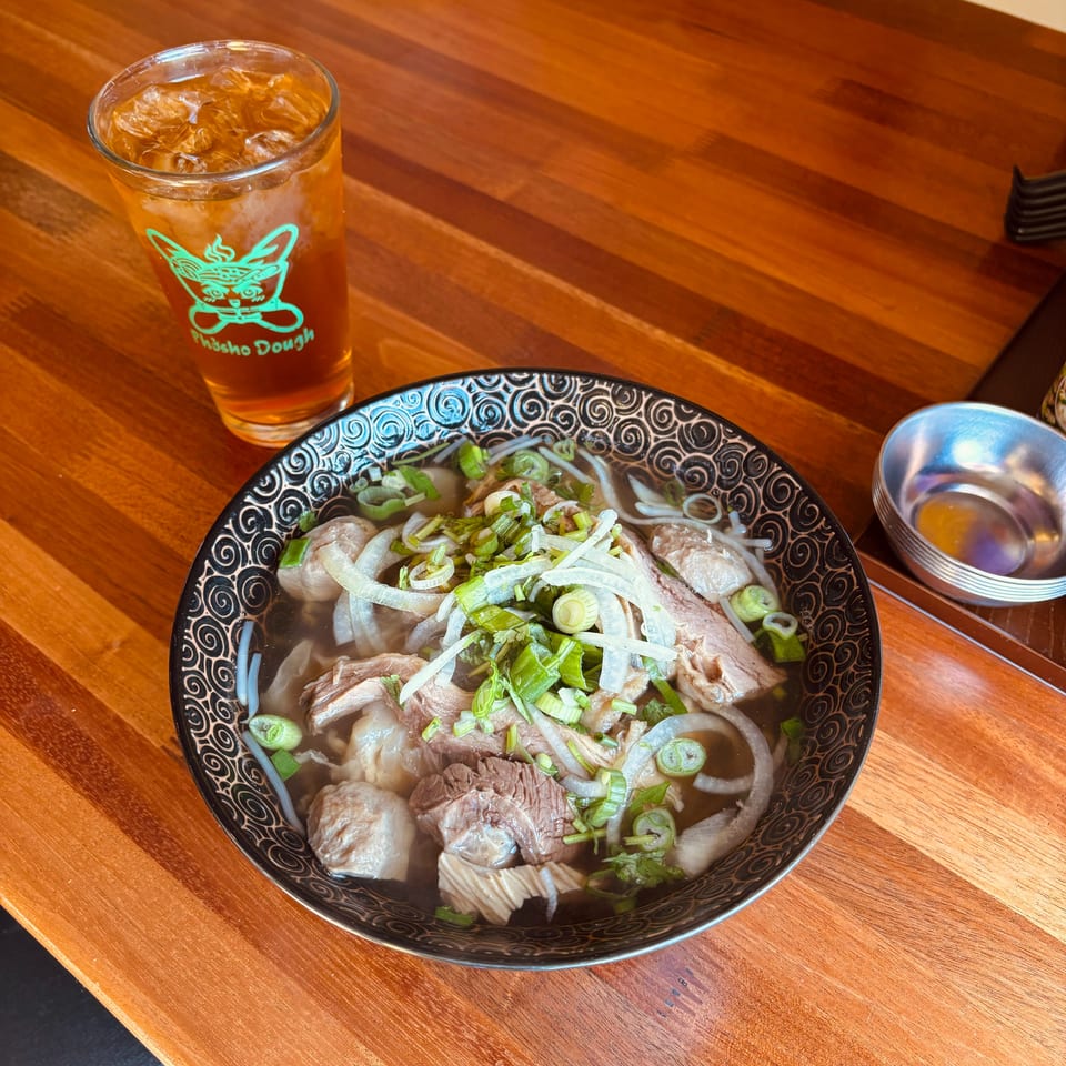 Bowl of noodle soup with sliced meat, meatballs, herbs, and scallions beside an iced amber drink on a wooden restaurant table.