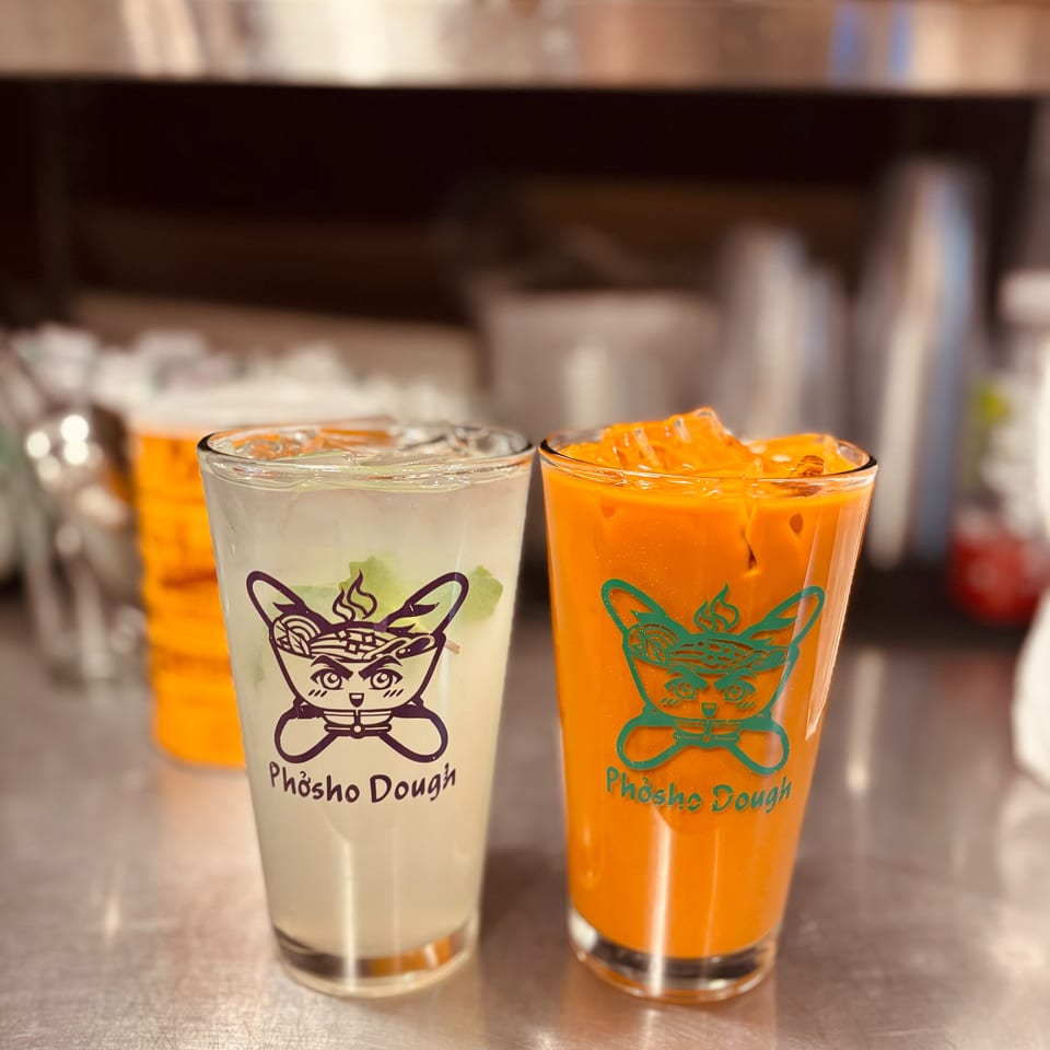 Two Phosho Dough branded drinks, one light and one orange, with ice on a metal counter.
