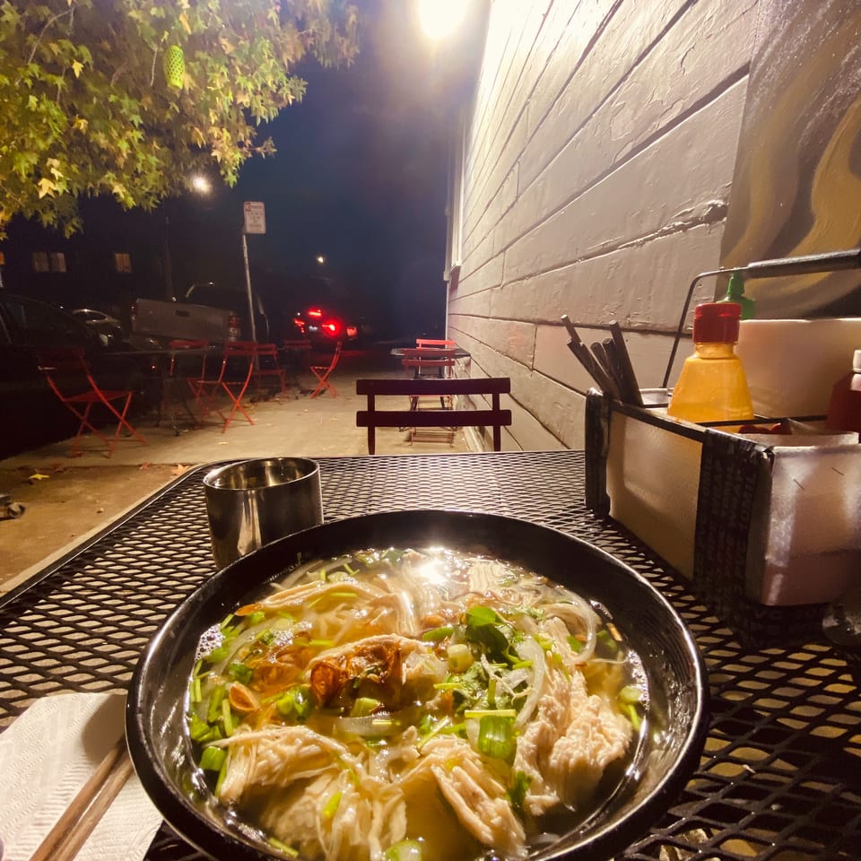 Chicken pho on a metal patio table at night, with outdoor seating and a building wall.