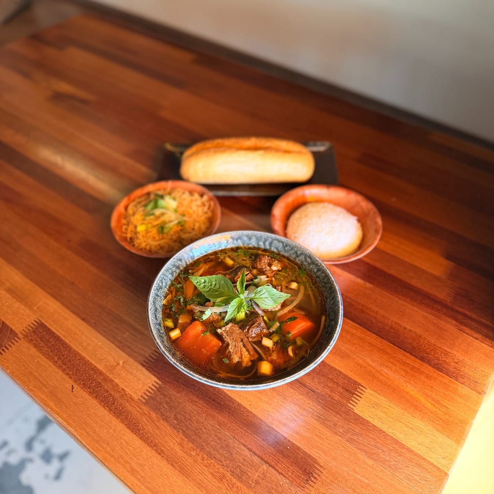 Hearty beef and vegetable soup with basil, served with baguette, rice, and noodles on a wooden table.