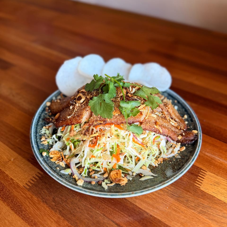 Crispy pork belly salad with shredded cabbage, carrots, cilantro, peanuts, and prawn crackers on a wooden table.