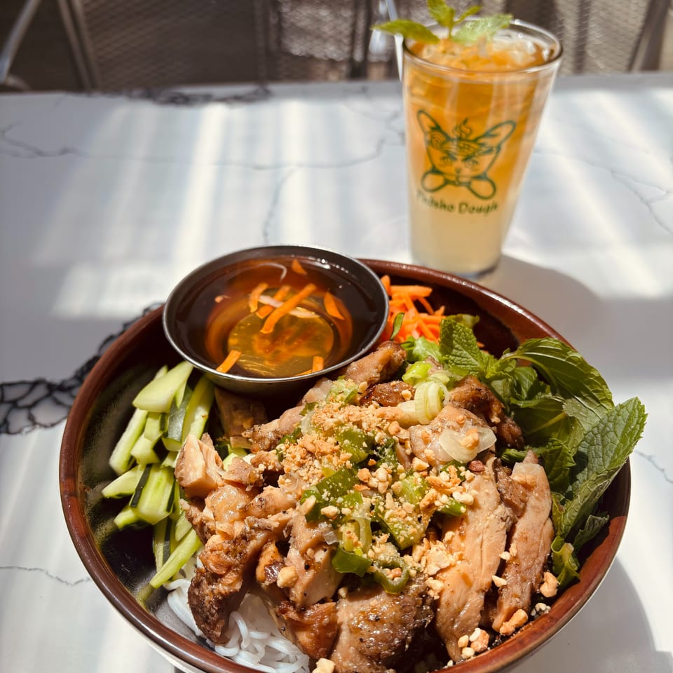 Vermicelli noodle bowl with grilled chicken, vegetables, peanuts, and sauce, beside an iced "Pudaho Dough" drink.