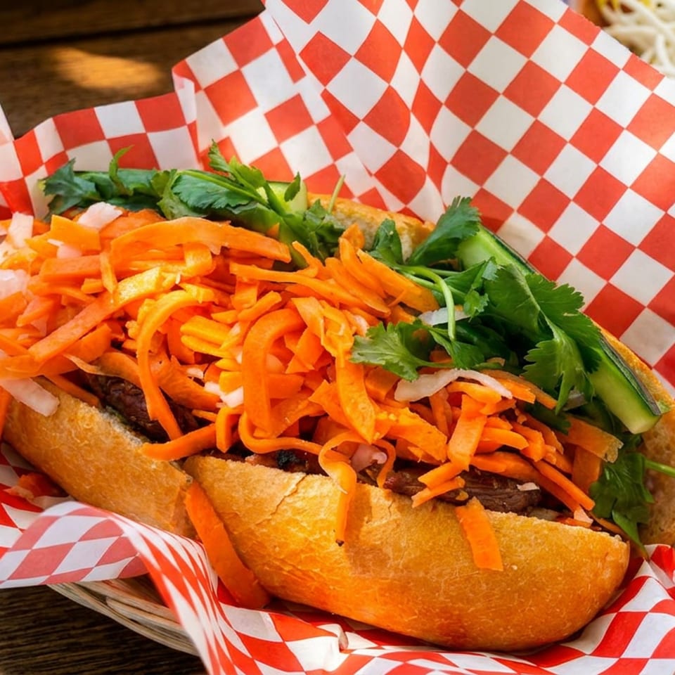 A banh mi sandwich with shredded carrots, cilantro, and cucumber in a red and white checkered paper.