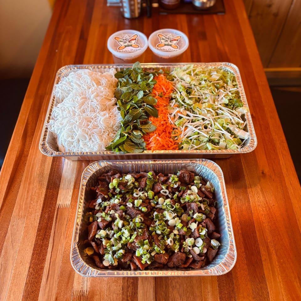 Two foil trays of Vietnamese food, one with noodles and vegetables, the other with meat and scallions.